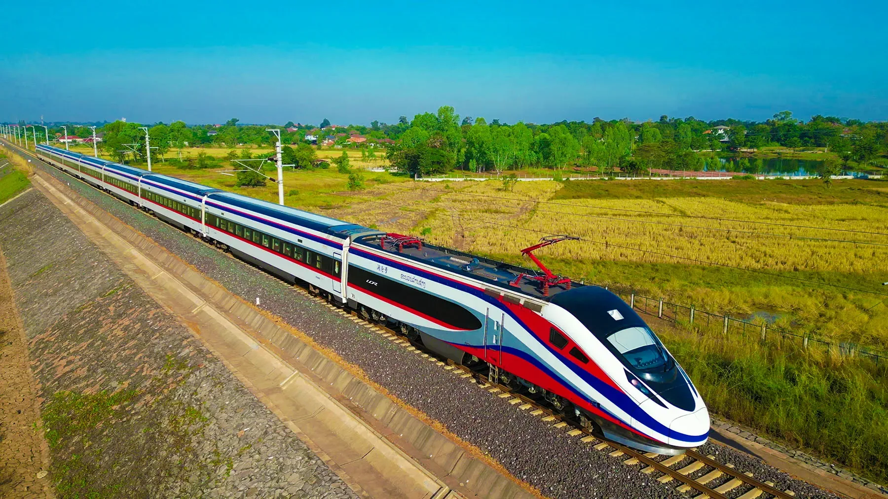 A Laos–China Railway train in red, white and blue livery crossing paddy fields